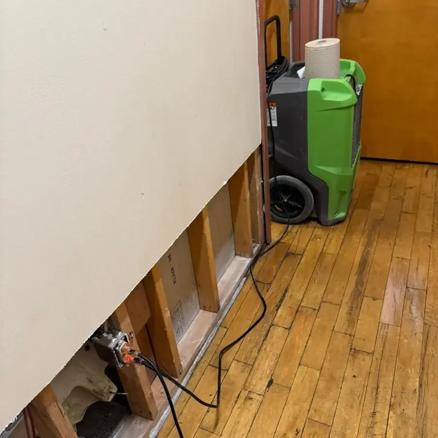 Dehumidifier beside an opened lower wall above hardwood flooring during interior water damage cleanup