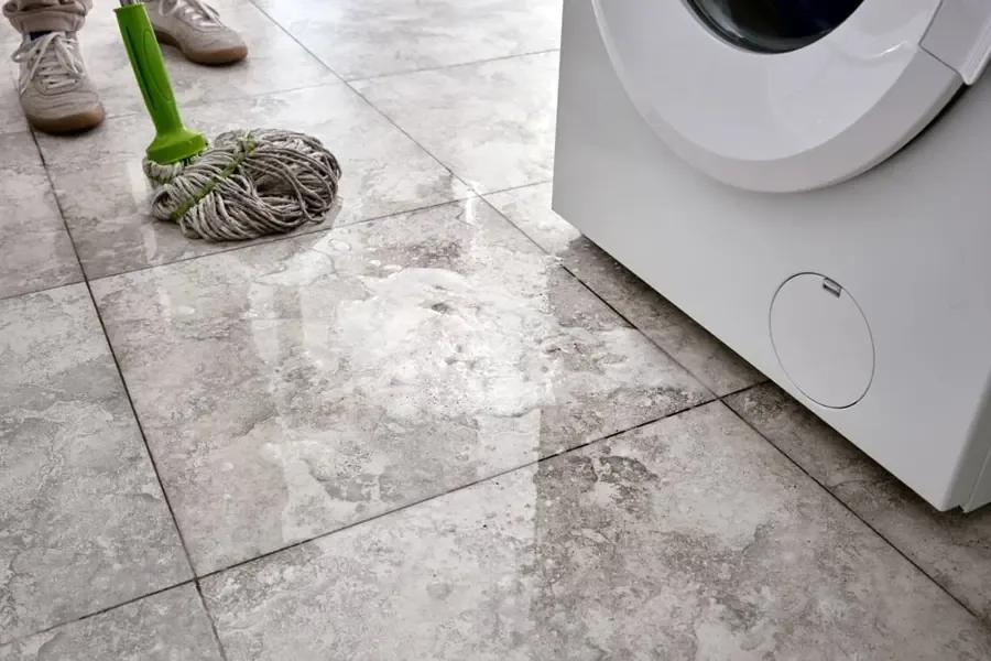 Water on a tiled laundry floor beside a washing machine and mop
